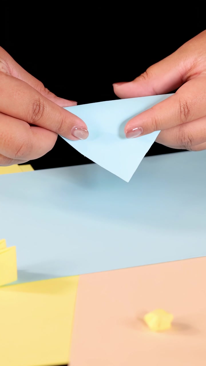 Una persona piega carta blu su un tavolo colorato, in primo piano, con illuminazione brillante e telecamera fissa dall'alto.