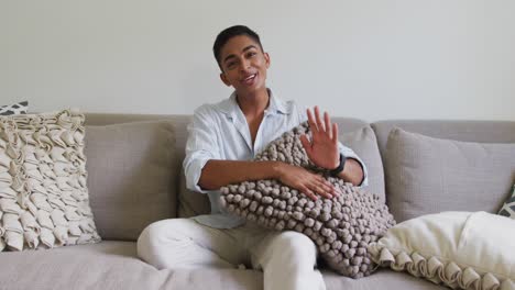 Happy-mixed-race-man-sitting-on-sofa-holding-cushion-laughing-and-waving
