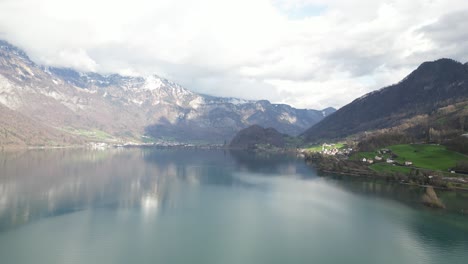 Rising-drone-clip-showing-calm-lake-with-green-grassy-shoreline-and-snowcapped-mountains