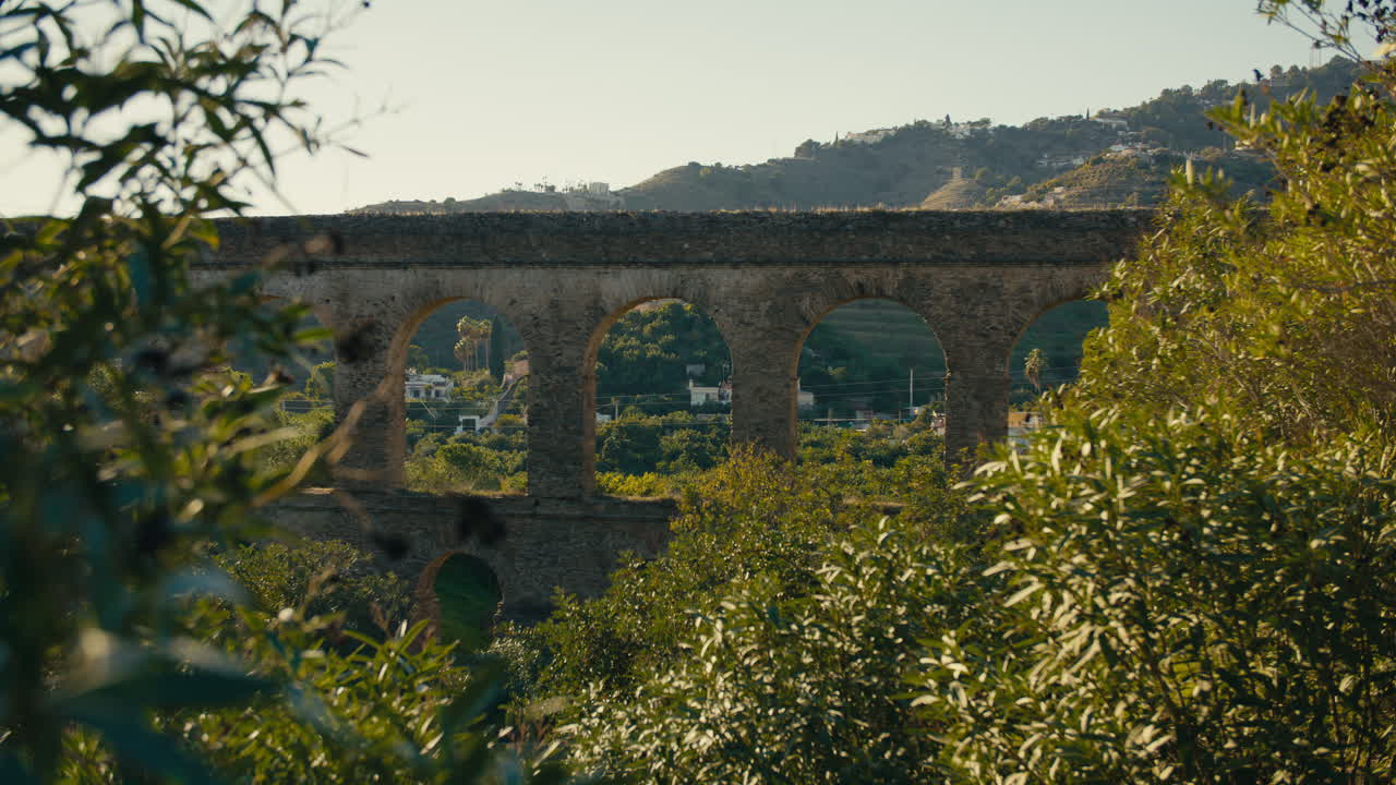 Ancient stone aqueduct framed by greenery, with arched windows revealing a Mediterranean valley and hills in soft evening light