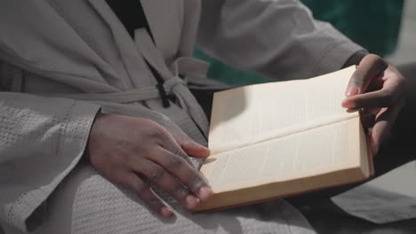 man in white robe holding open book, reading calmly while sitting on floor in peaceful bedroom. soft sunlight filters through window, creating serene, relaxed atmosphere for quiet reflection