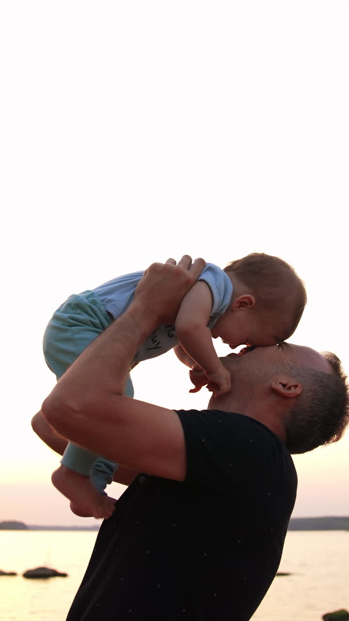 Loving dad talking to and kissing his little son. Laughing baby boy stepping his bare feet by father's chest. River at backdrop. Vertical video