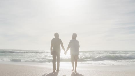 Vista-Posterior-De-Una-Pareja-De-Ancianos-Hispanos-Tomados-De-La-Mano,-Caminando-Por-La-Playa-Al-Atardecer