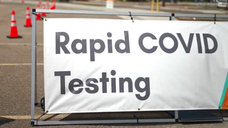 rapid covid 19 testing site sign posted outside on sunny summer day in empty parking lot with orange cones setup, and cars driving by in background