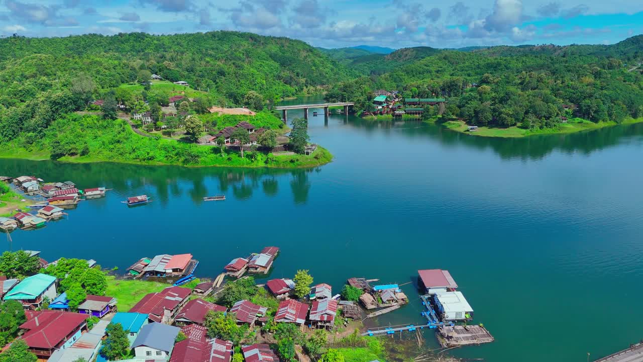 Drone pushes in over Sangkhlaburi, Thailand. Aerial view of floating houses, river, bridge, lush green hills, and blue sky with scattered clouds