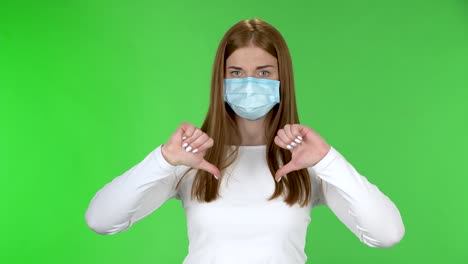 portrait of young girl in medical protective face mask looking at camera and disgruntled shakes his head and shows fingers down on both hands