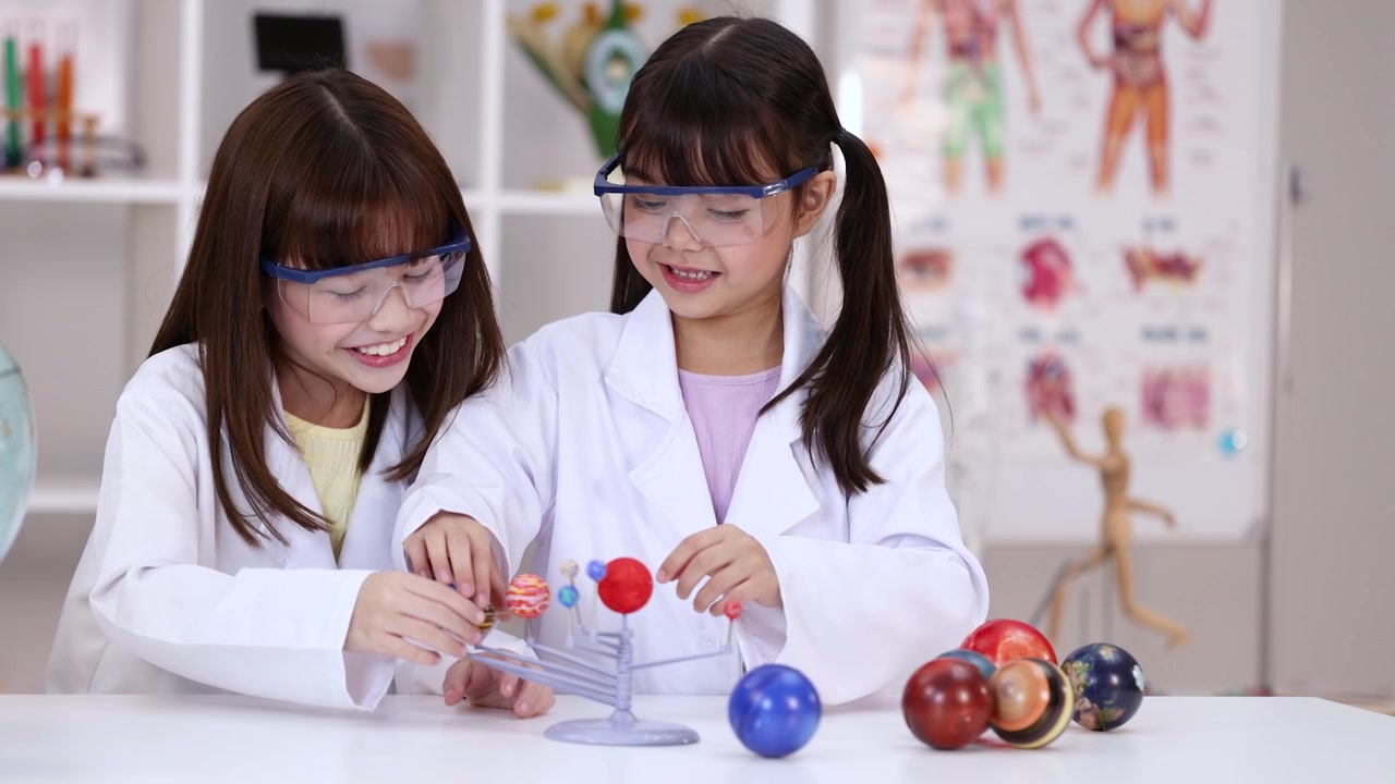 Two girls in lab coats and goggles examine a solar system model in science classroom.