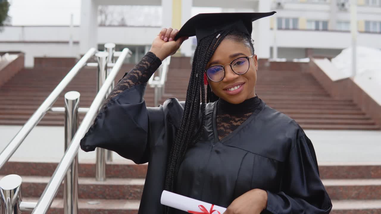 una mujer joven en la universidad en el manto del maestro sostiene un diploma con una mano y un sombrero con otra. maestro de ciencias mira a la cámara. maestro de farmacia