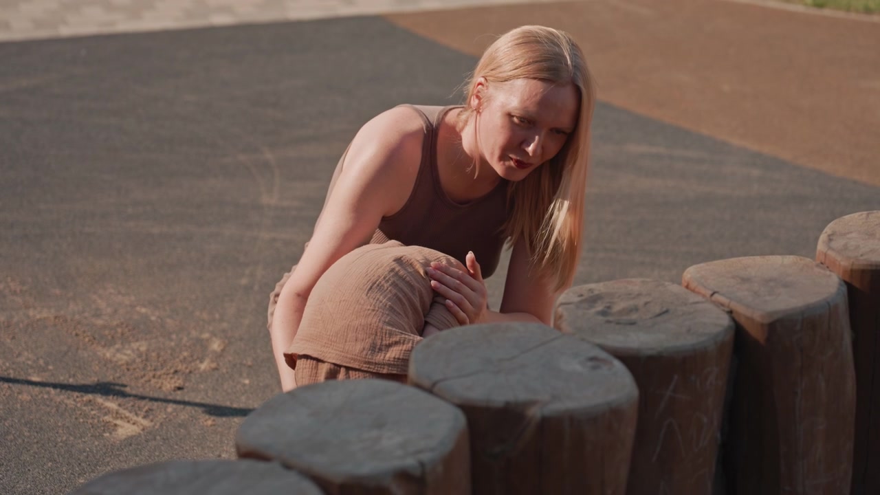 Una donna è seduta a terra accanto a tronchi di legno in un ambiente esterno, apparendo contemplativa o a riposo. La donna è vestita in modo casual con capelli lunghi e un top senza maniche, circondata da luce naturale.