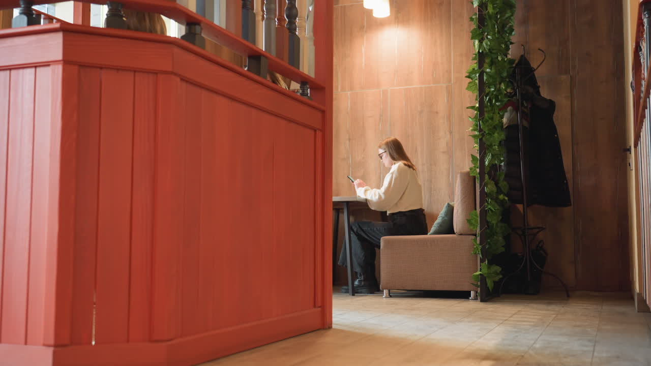 Young woman seated alone under warm overhead light operating phone in cozy cafe, surrounded by wooden wall panels, coat hanger with clothes, leafy decor, and modern equipment in background
