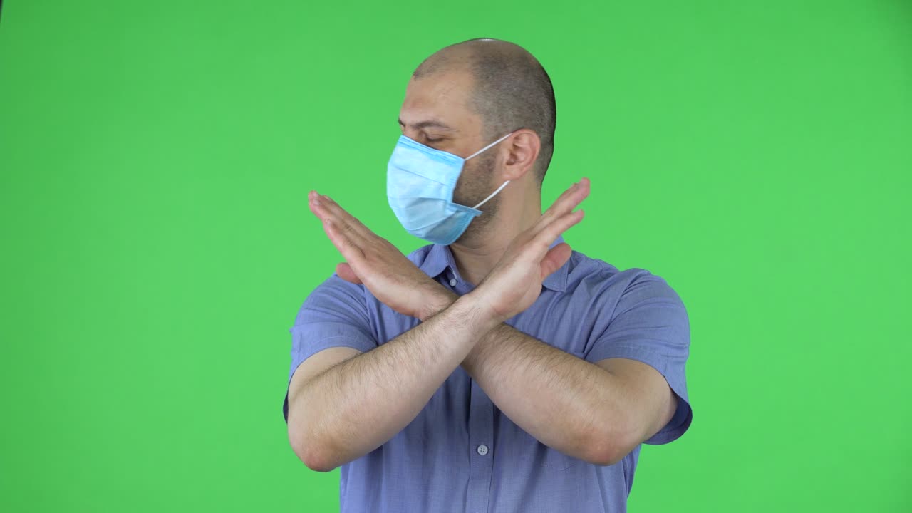 retrato de un hombre de mediana edad con máscara médica muestra un gesto de brazos cruzados que significa no. hombre calvo en camisa azul posando en pantalla verde en el estudio.