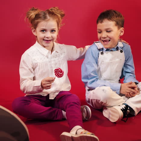 smiling little children playing with tasty lollipop over red background. funny boy and happy girl sitting on the floor together among beautiful decorations.