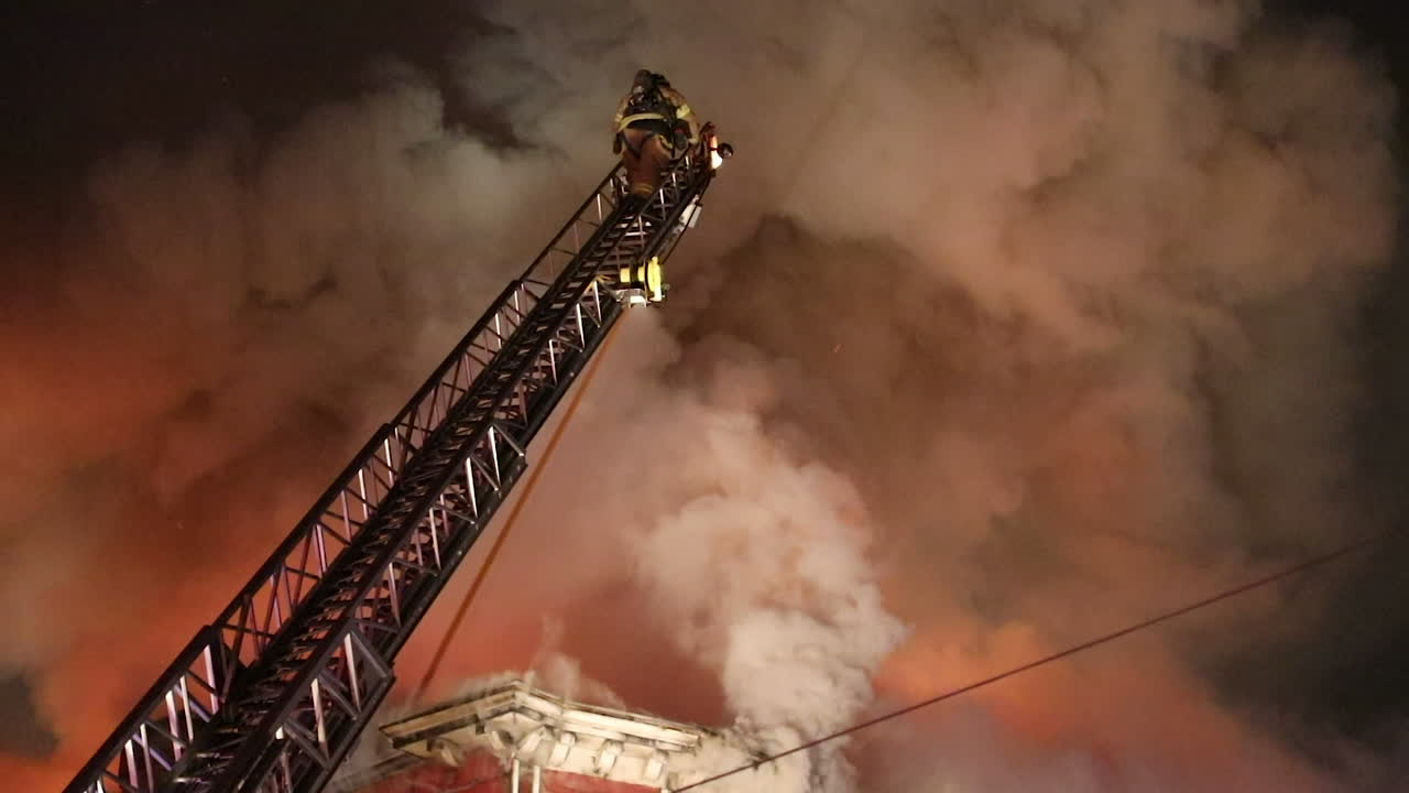 A Brave Firefighter Climbs a Ladder Towards Massive Plumes of Smoke and Flames Coming out of a Large Apartment Building at Night