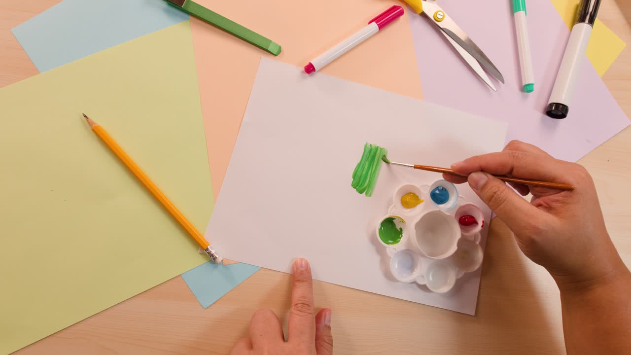 Artist paints green brushstrokes on blank paper with palette, overhead view, soft even lighting