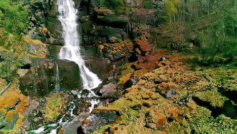 Drone-shot-pulling-back-to-reveal-a-large-waterfall-in-Norway,-two-people-standing-underneath-it