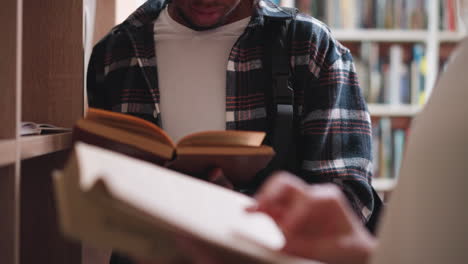 students positioned by bookshelves in library. black man and white woman flip pages searching for literature to prepare for college exams. team education