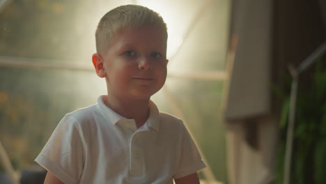 kid smiles and looks at camera standing in front of lighted transparent film of tent. cute blond preschooler enjoys evening outdoors at glamping site closeup on blurred background