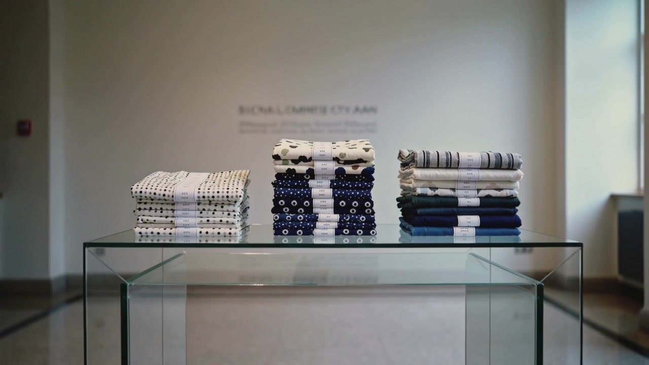 Pulling back camera revealing three stacked textiles on glass table at gallery, showing white bands