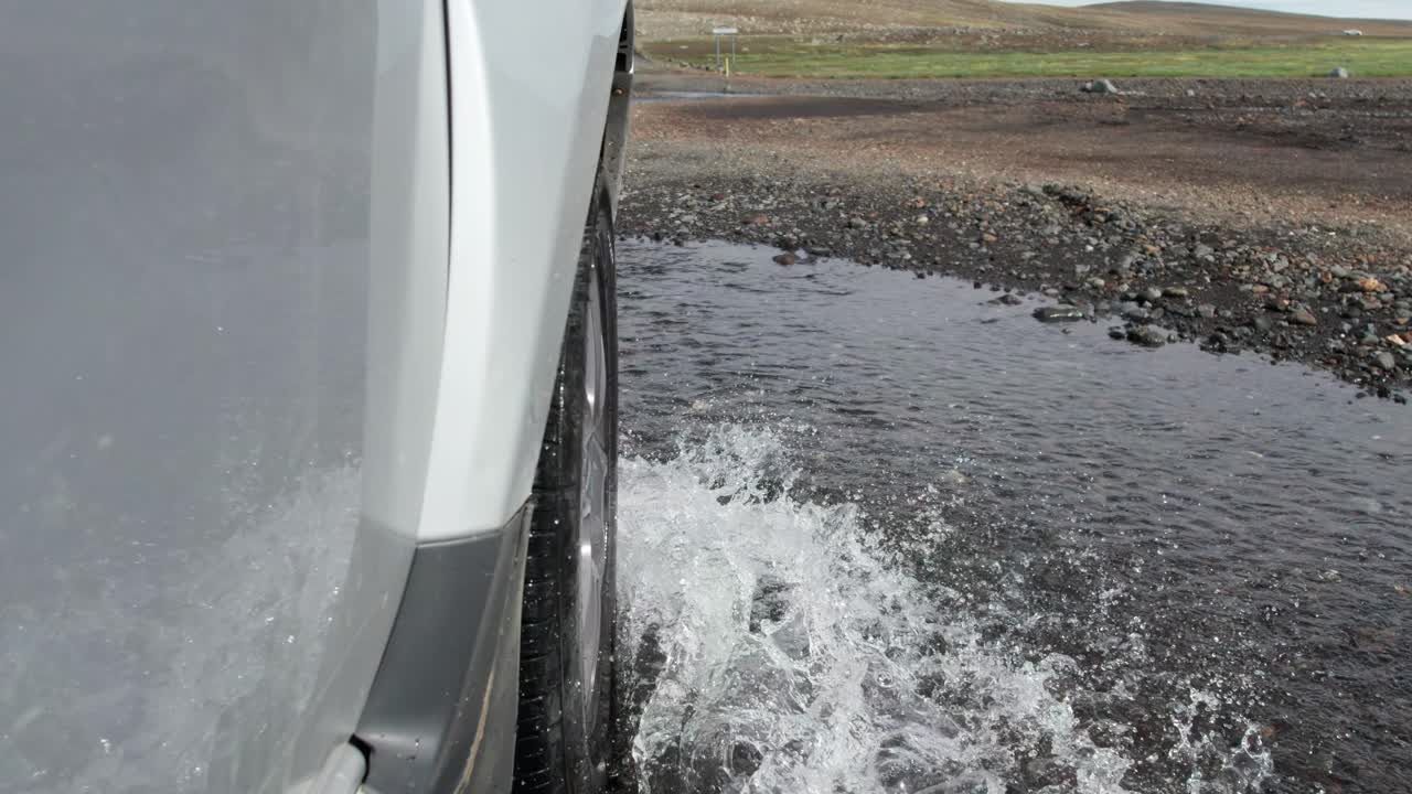 A close-up shot of a car driving through a flooded river on remote mountain roads in Iceland. The vehicle splashes through the water as it navigates the rugged and challenging terrain.