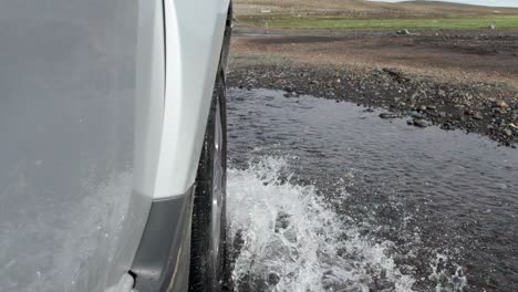 a close-up shot of a car driving through a flooded river on remote mountain roads in iceland. the vehicle splashes through the water as it navigates the rugged and challenging terrain.