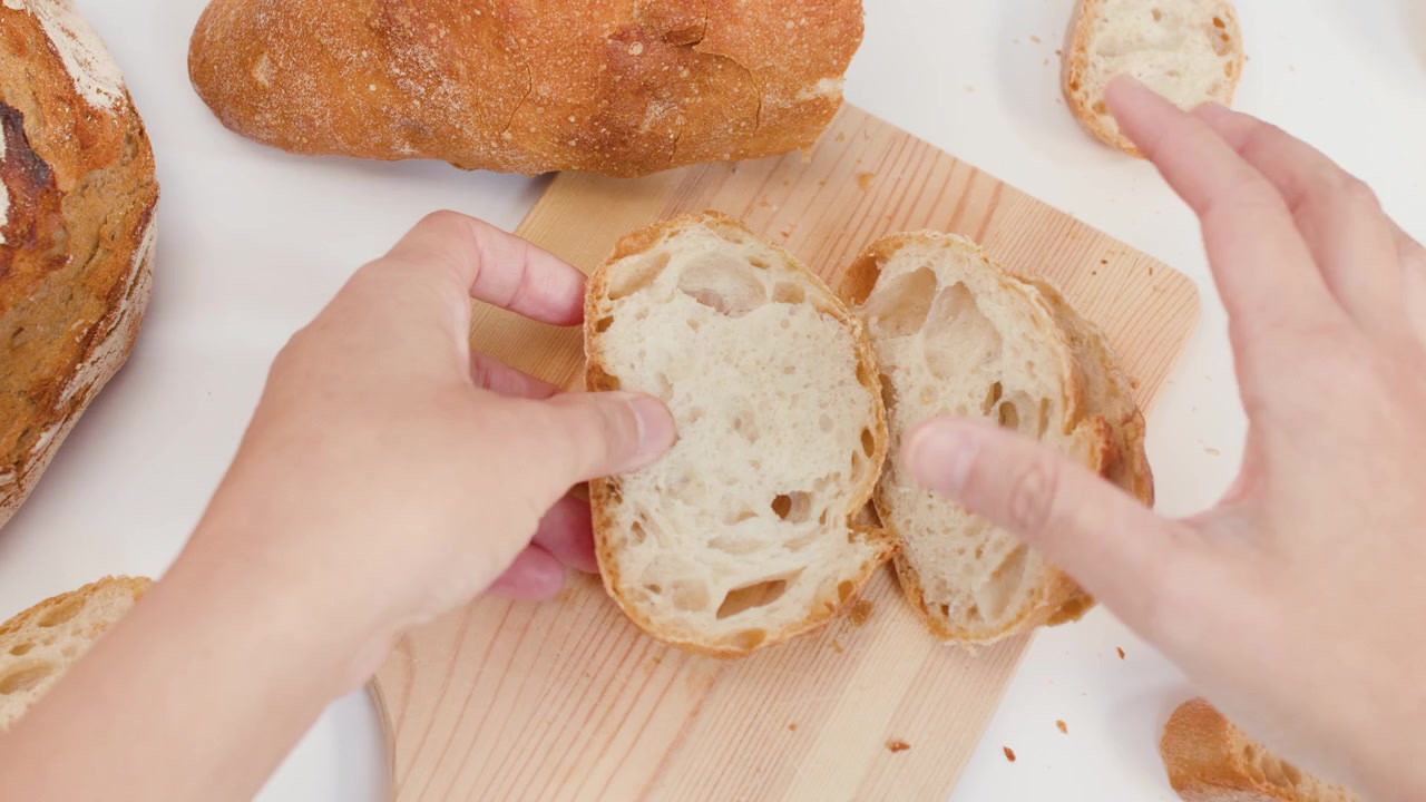 Top-down view of hands organizing fresh crusty bread slices on a light wooden board.