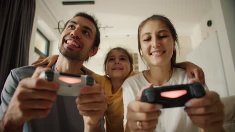 happy young family playing video games using joysticks in a modern apartment. a brunette man with a gray t-shirt plays video games with his wife, a brunette girl in a white t-shirt, using a joystick, and their little daughter in a yellow dress hugs her parents and is happy for them in a modern room