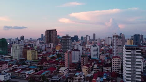 Phnom-Penh-city-as-the-sun-sets,-viewed-by-drone,-level-with-the-high-rise-buildings-and-impressive-sky-in-the-background,-camera-panning-around-to-the-right