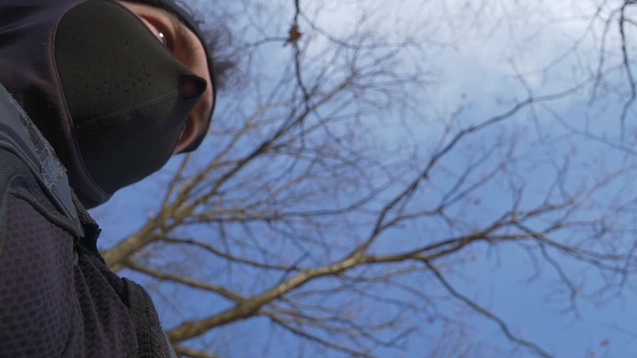 Low-angle shot of a masked individual in winter gear looking up at bare trees under a clear blue sky. Ideal for concepts of solitude, mystery, or outdoor survival.
