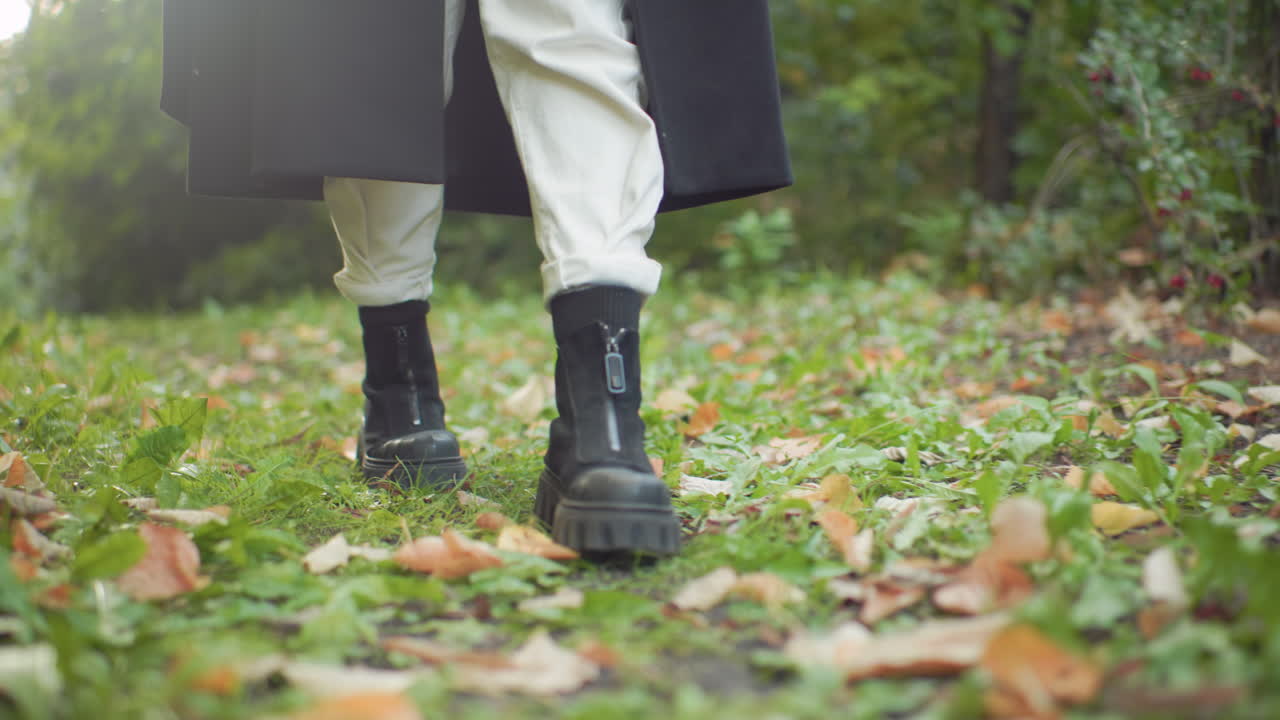 Leg view of casual woman in black boots walking on leaf carpet along green forest path, coat swinging over white trousers, slow steps through damp grass and scattered autumn foliage