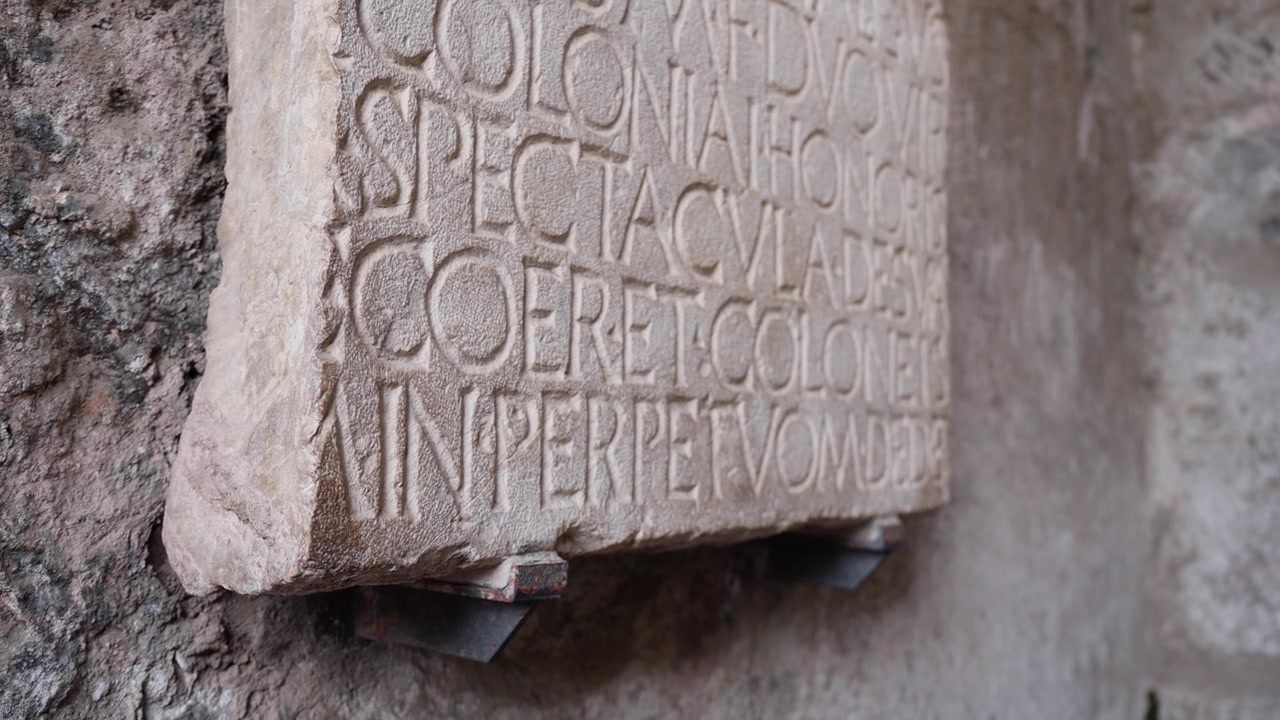 Antica pietra con iscrizione latina nelle rovine di Pompei, che mostra lettere romane incise su una lastra consumata dal tempo, probabilmente legata a spettacoli pubblici o a una dedica civica.