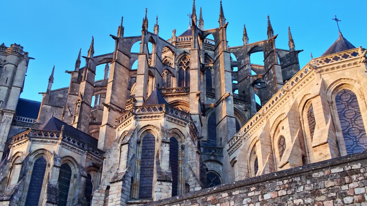 Plano panorâmico em ângulo baixo da Catedral de Saint-Julien em Le Mans sob um céu azul claro, destacando os arcos góticos, contrafortes e detalhes em pedra. França.
