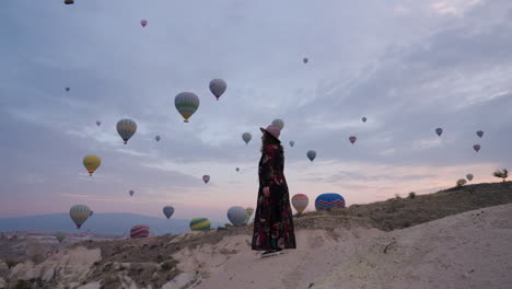 Mujer-Con-Vestido-Largo-Parada-En-Una-Montaña-Y-Mirando-Coloridos-Globos-Aerostáticos-En-Vuelo-En-Capadocia,-Turquía-Al-Amanecer