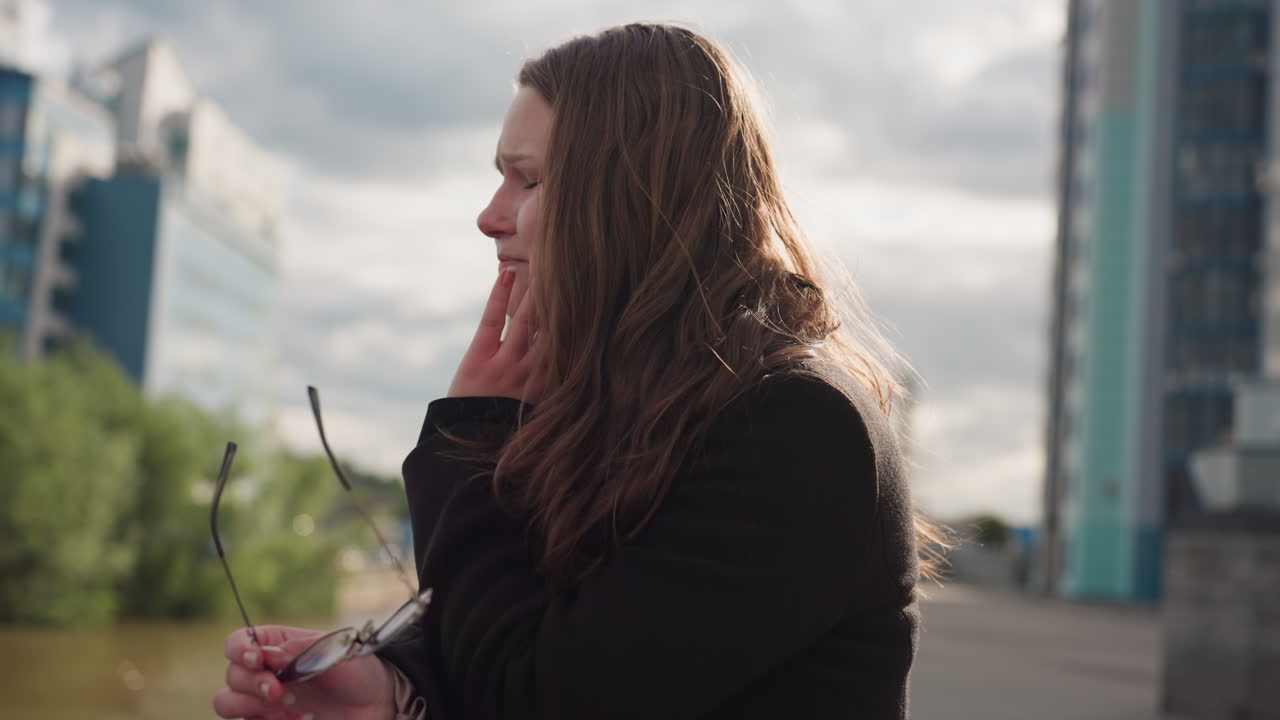 Tourist wearing thick jacket covers face in discomfort while standing by river, sunlight reflecting on glasses as skin turns red, expressing sensitivity and tension amid calm modern architecture