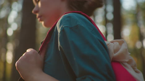 close-up of woman in green shirt holding red bag with beige cloth draped over it, walking in forest as sunlight reflects softly on her with background blur featuring trees