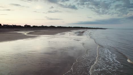 Von-Oben-Zeigen-Luftaufnahmen-Einen-Bezaubernden-Sonnenuntergangsstrand-Mit-Schimmerndem,-Nassem-Sand,-Einem-Ruhigen-Lila-rosafarbenen-Meer-Und-Menschen,-Die-Mit-Der-Silhouette-Ihres-Hundes-Spazieren-Gehen