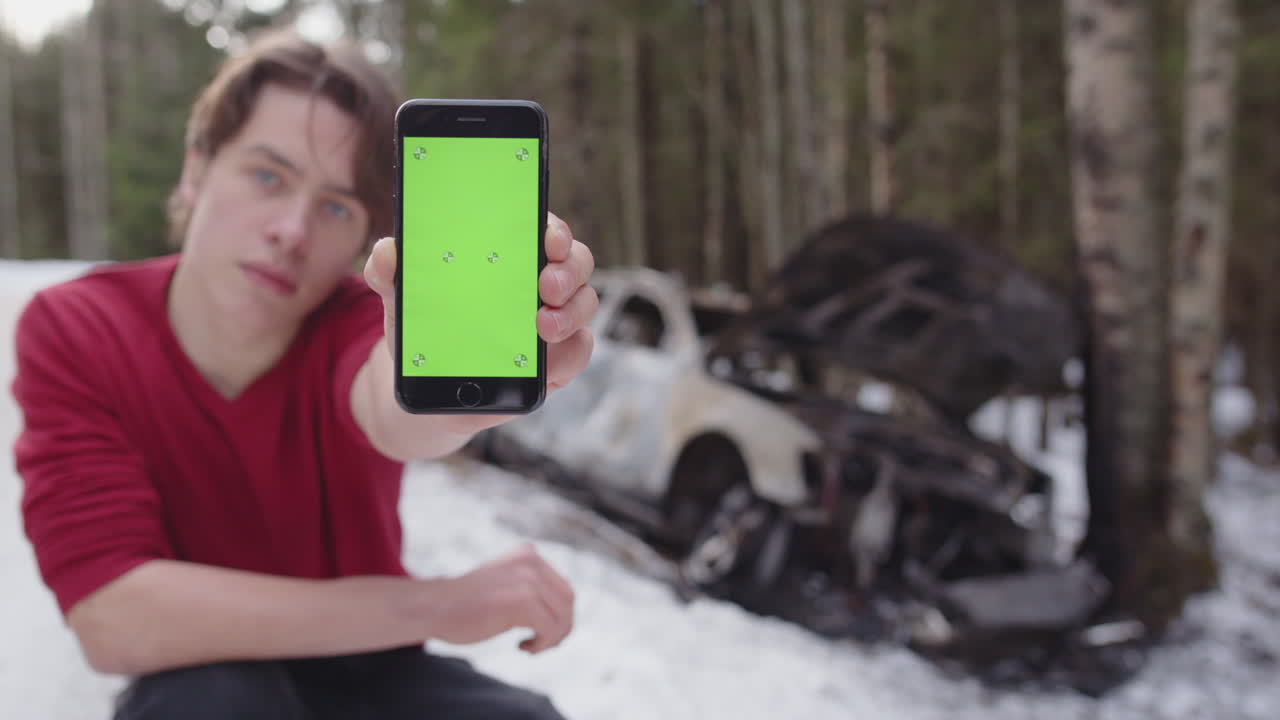 Crouching student looks back at wrecked car holds out phone with green screen