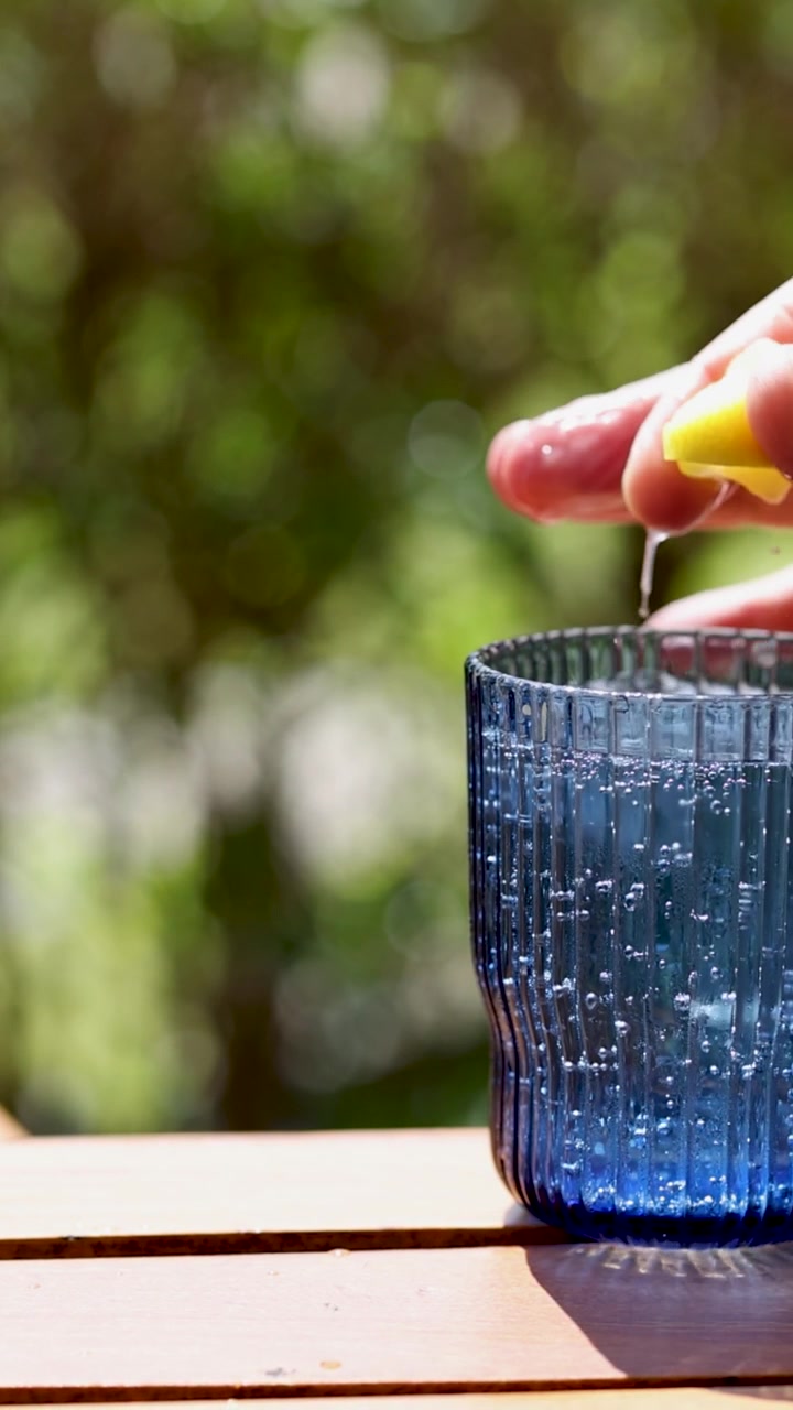 A hand squeezes lemon into a blue glass on a sunlit table, creating a refreshing scene