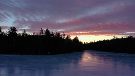 aerial slide over a frozen pond with the forest silhouetted against a pink and yellow sunrise