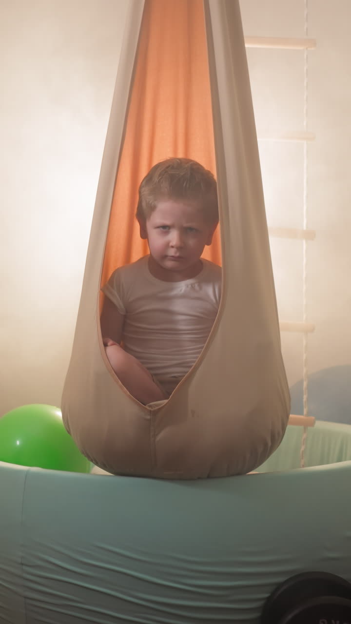 Little boy frowns sitting in orange hammock hanging above ball pit. Cute blond toddler relaxing in modern flat gym with training equipment slow motion