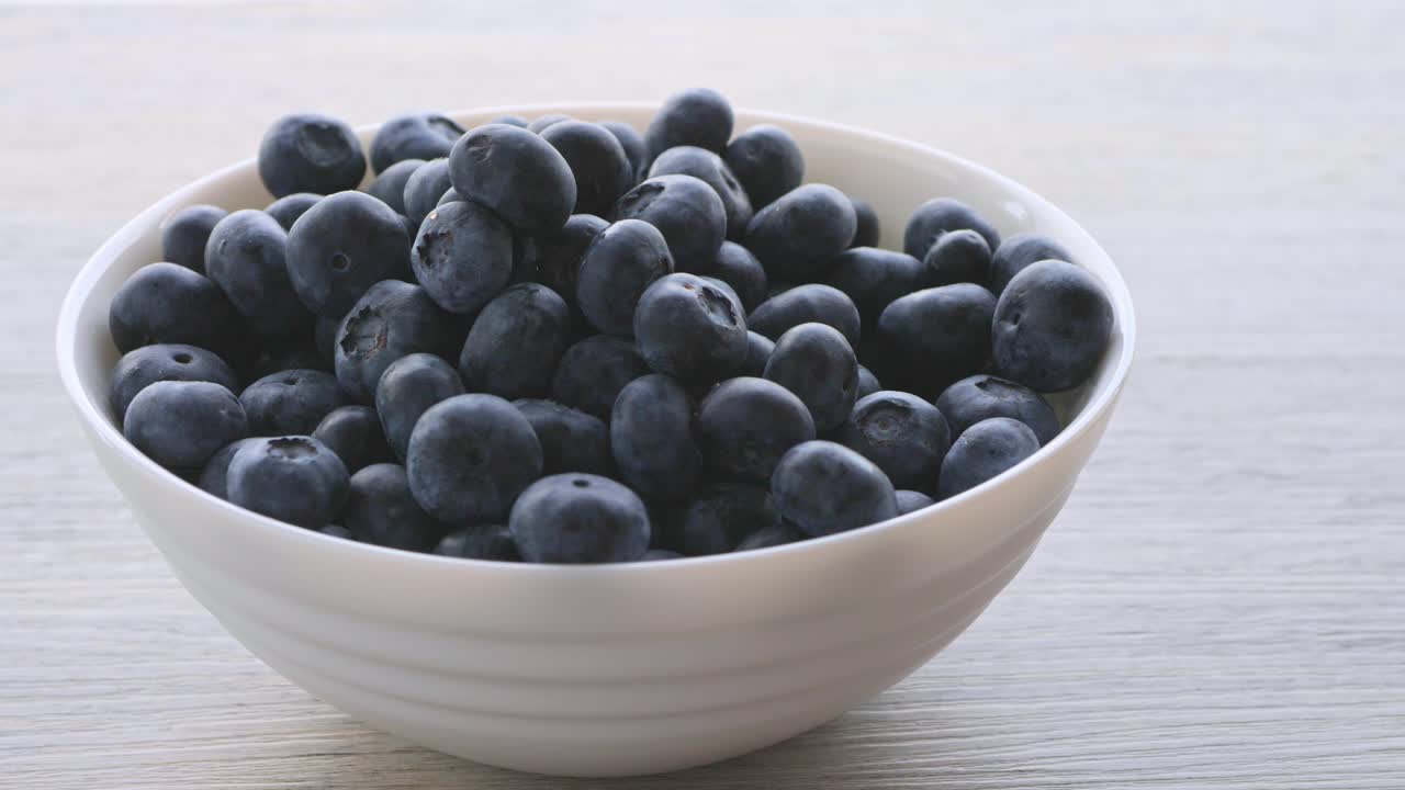 Fresh blueberries falling into a white bowl on a light wooden table, healthy organic fruit concept
