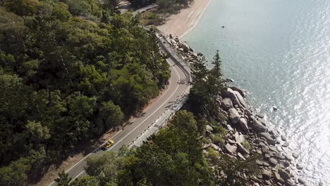 aerial view of open top convertible classic beach buggy driving along coastal clifftop road next to with sun reflecting off crystal clear tropical water