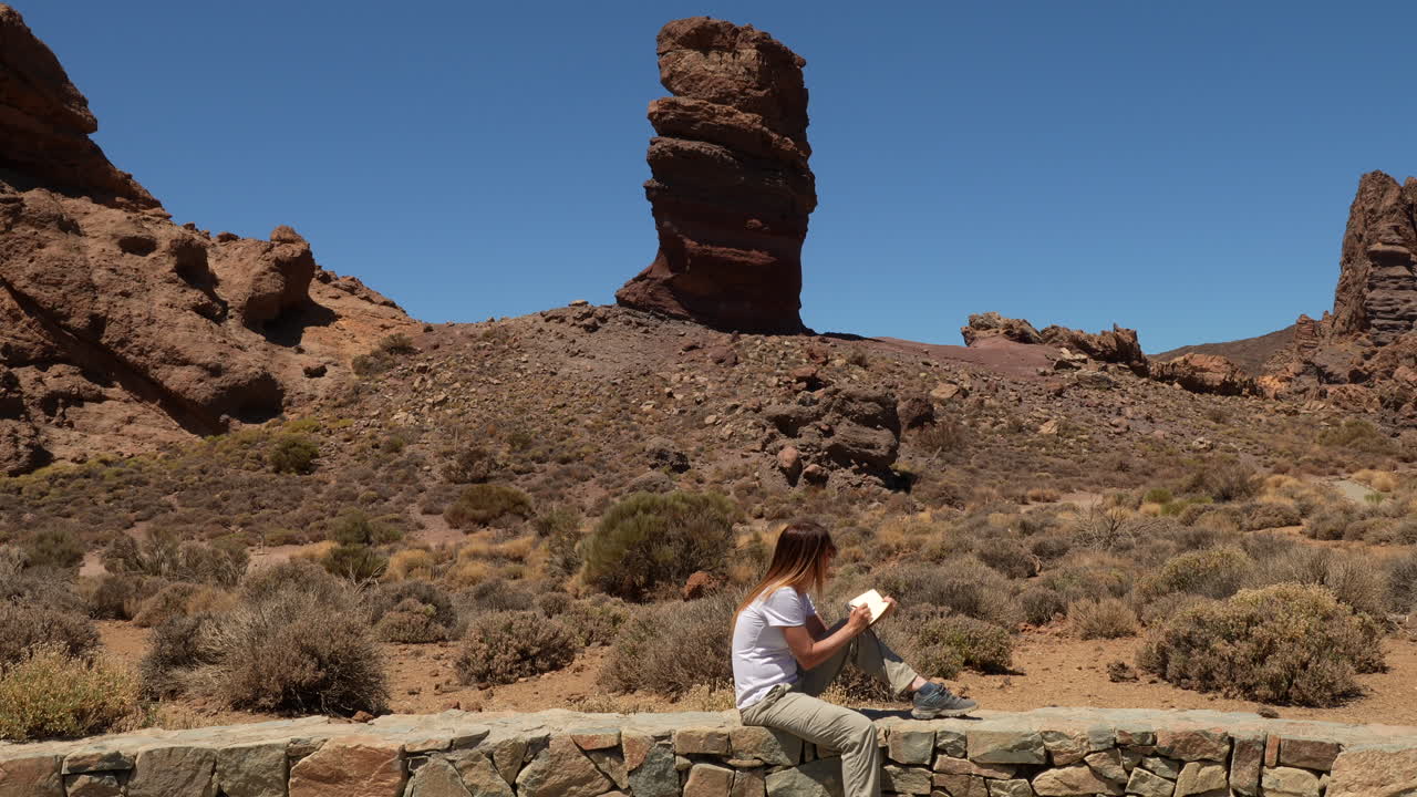 Female traveler sitting on a stone wall and reading a book with the Roque Cinchado rock formation in the background in Spain