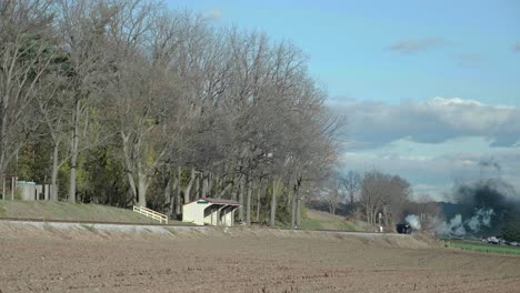 in a tranquil countryside, smoke billows from a fire in the distance while trees line a path. a small shelter stands nearby, capturing the serenity of the setting.