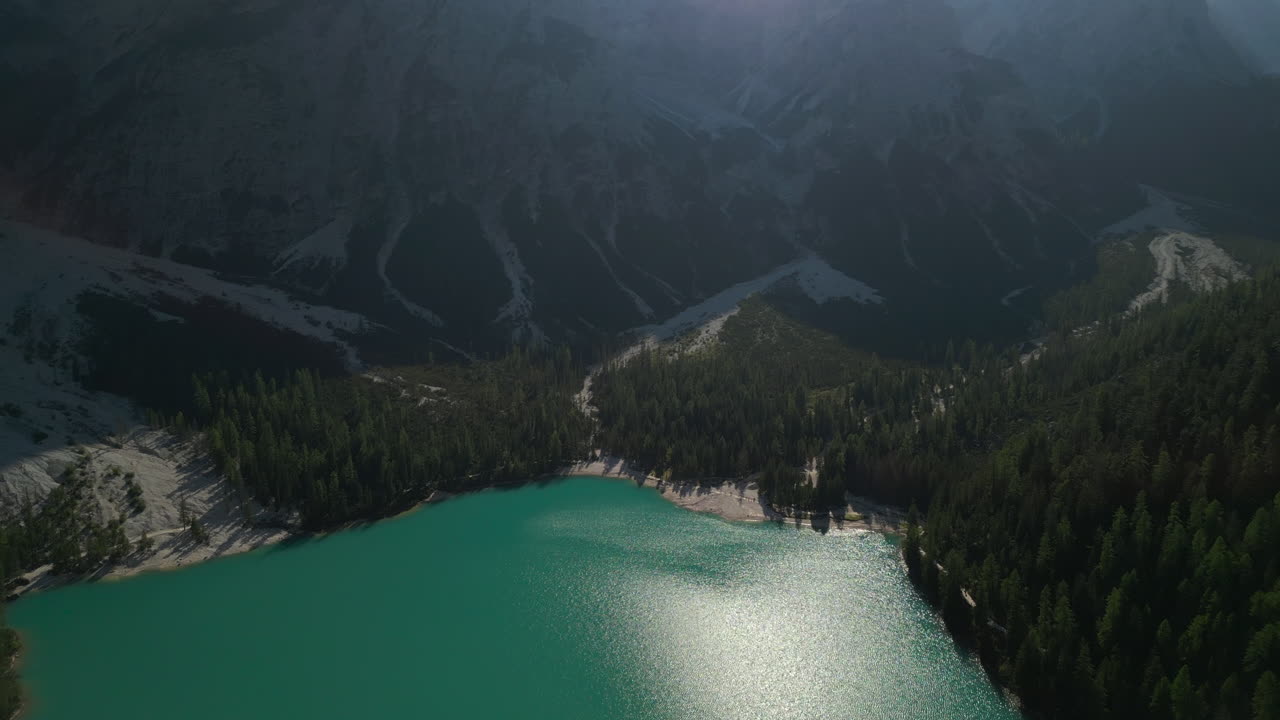 Passaggio su acque verde smeraldo che si avvicinano alla riva del lago e a un'imponente montagna rocciosa. I raggi del sole si riflettono sulla superficie tra profonde ombre mattutine. Ripresa aerea frontale ad alta quota in 4K, Apple ProRes a 25fps