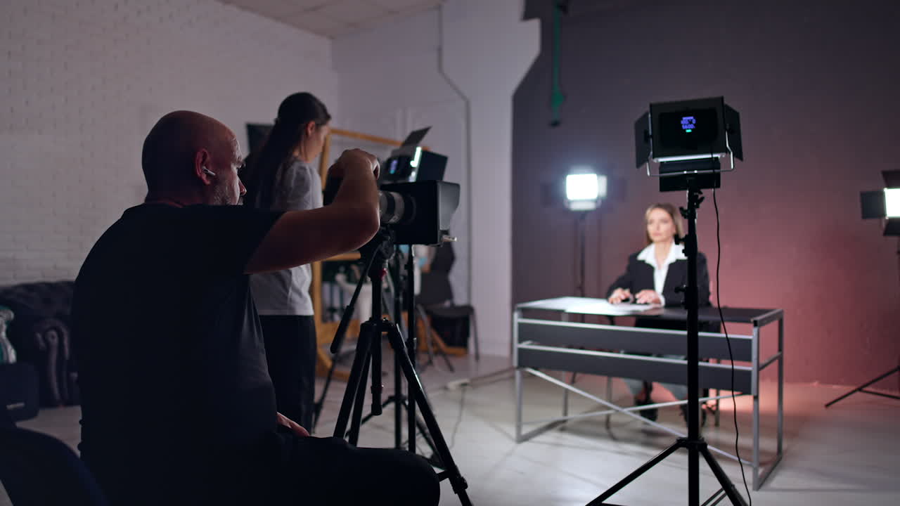 Video crew working in photo studio creating blog content. Female blogger sitting at desk talks to camera. Man taking pictures.