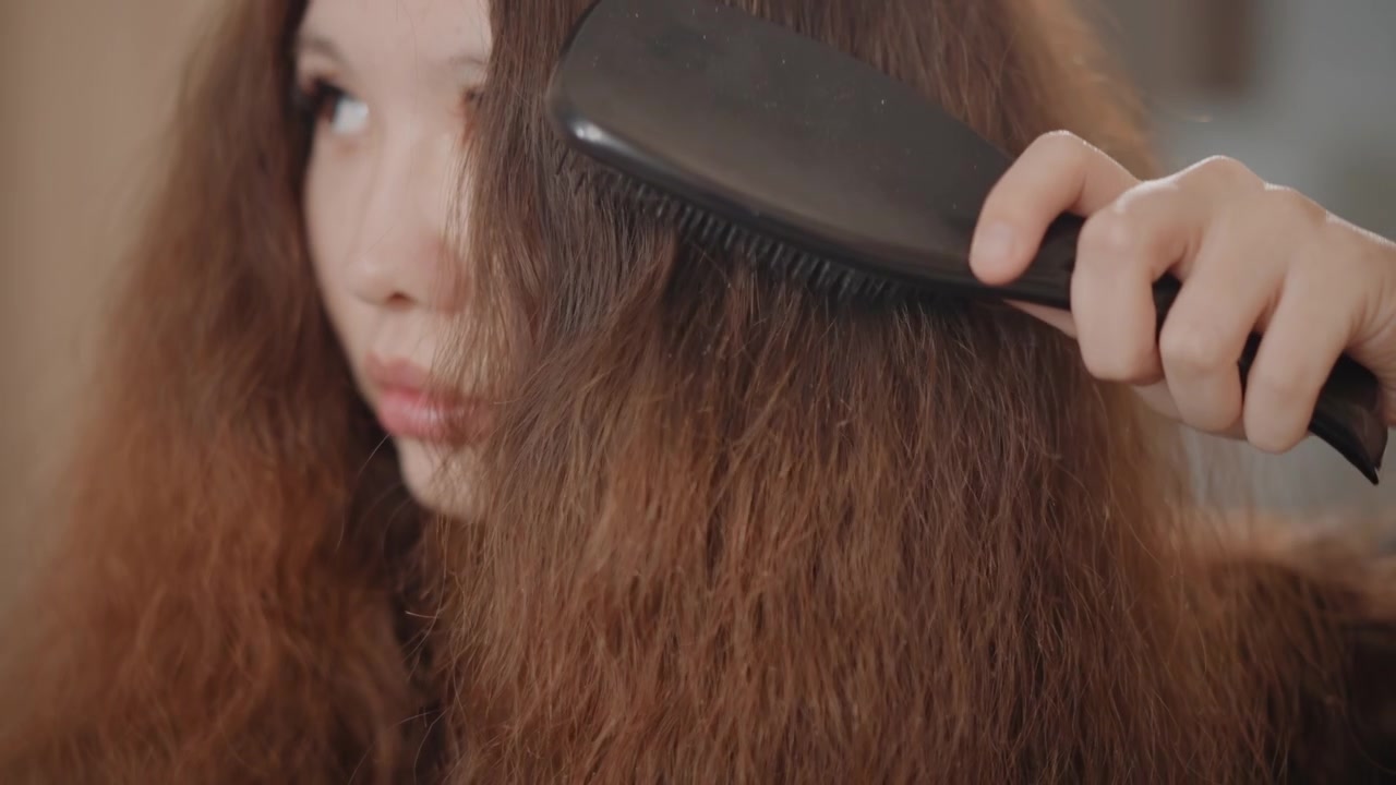 Potret close-up setinggi mata seorang wanita muda dengan rambut cokelat tebal dan mengembang yang sedang disisir dengan sikat dayung lebar; tangan mengangkat sikat melalui rambut tebal, menyebabkan helai rambut terpisah, terangkat, dan bergerak.