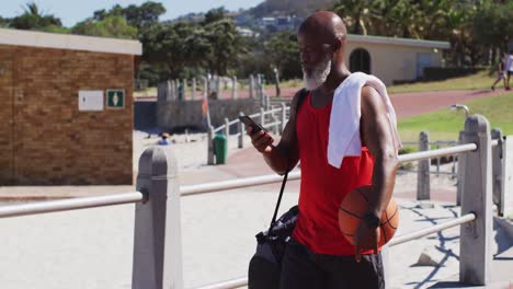 senior african american man with basketball talking on smartphone while walking near the beach