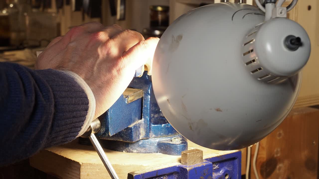 Close-up of a luthier adjusting a lamp while carving a violin wooden scroll held in a bench vise
