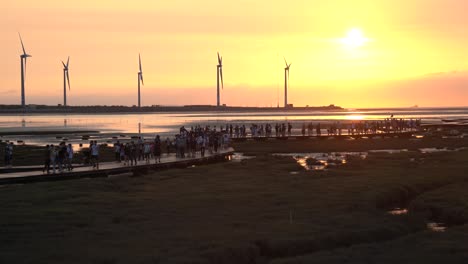 Vibrant-orange-sunset-landscape-shot-of-tourists-strolling-on-wooden-walkway-platform-at-beautiful-Gaomei-wetlands-preservation-area-with-wind-power-plant-in-the-background-in-Taichung,-Taiwan