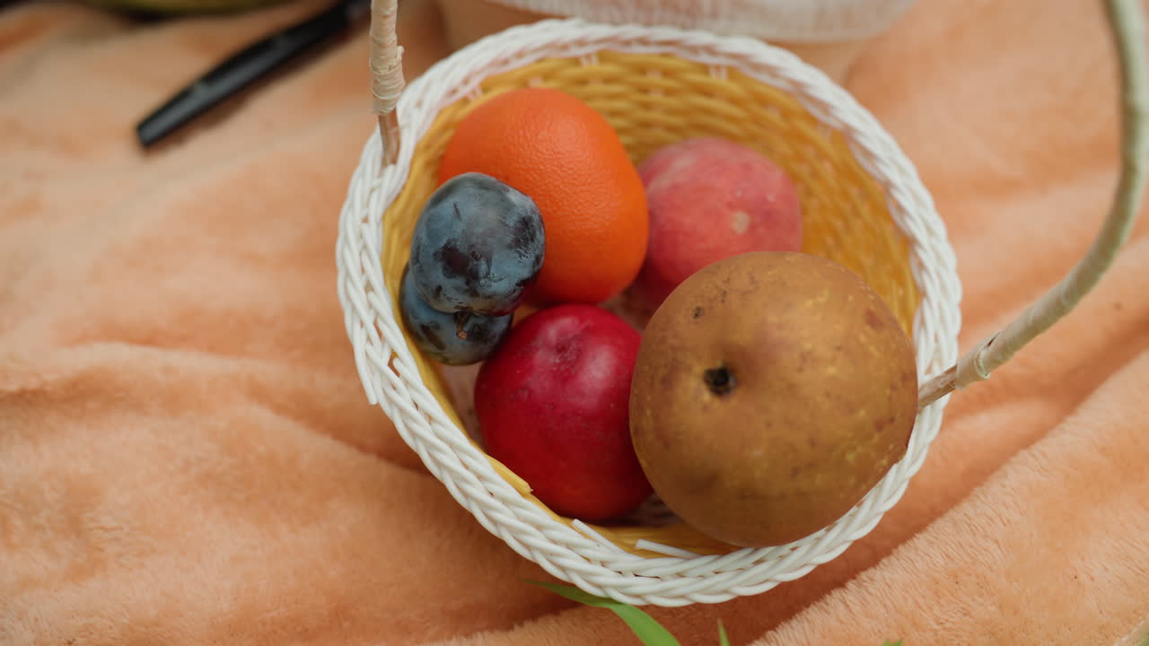 kurv med blandet frukt på teppe, intim piknikscene med flettet kurv som holder oransje, rødt eple, kiwi og lilla plomme, nærbilde av manikyrert hånd som forsiktig velger frukt, varm plysjbakgrunn, myk